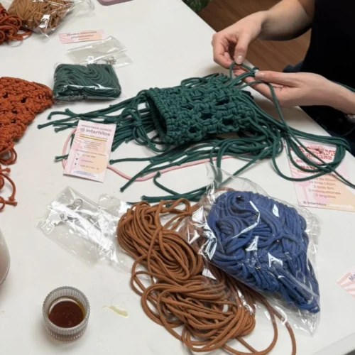 Persona tejiendo un phone holder verde en un taller, con hilos de colores y materiales textiles de Interhilos sobre la mesa.